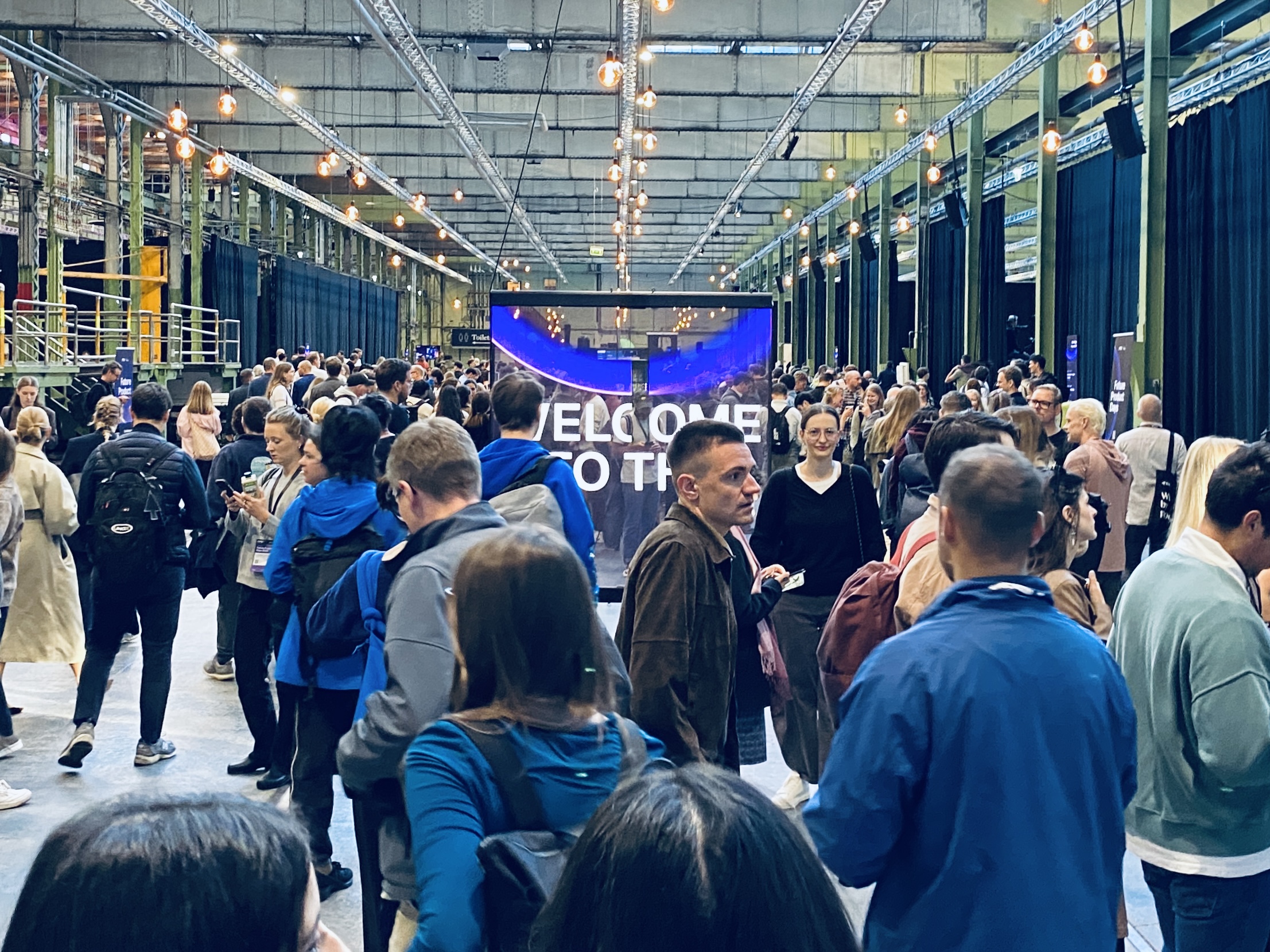 Image of a big hall with lots of people walking across each other and in the middle the billboard that says 'Welcome to the Future of Product 2026'